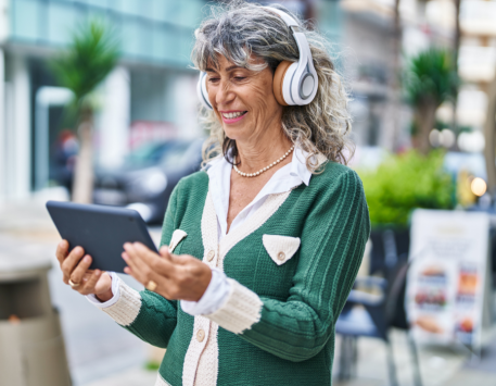Mid 50s female with grey curly hair, walking outside with headphones on and smiling at tablet